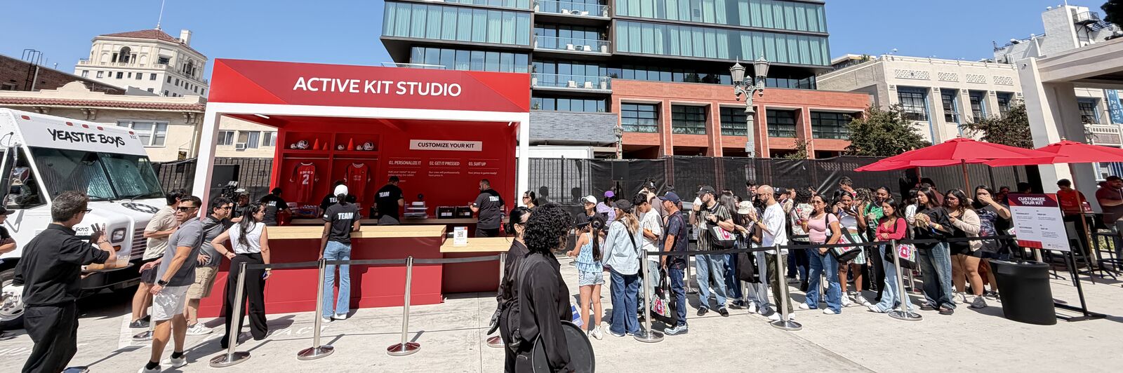 Red outdoor jersey personalization booth on a city plaza, staffed crew at the counter and a long stanchioned guest queue stretching across the concrete