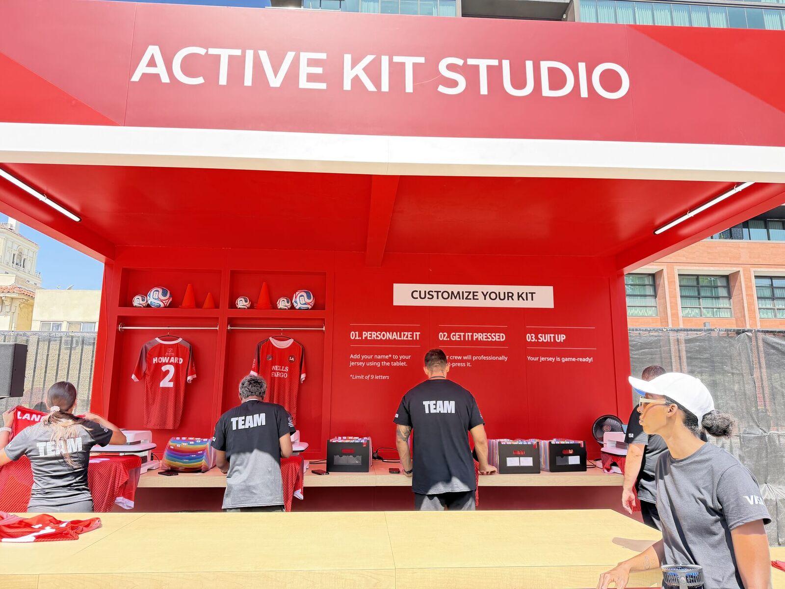 Red outdoor jersey kit studio with tablet entry stations on the left and a pressing crew in TEAM shirts working the counter behind them